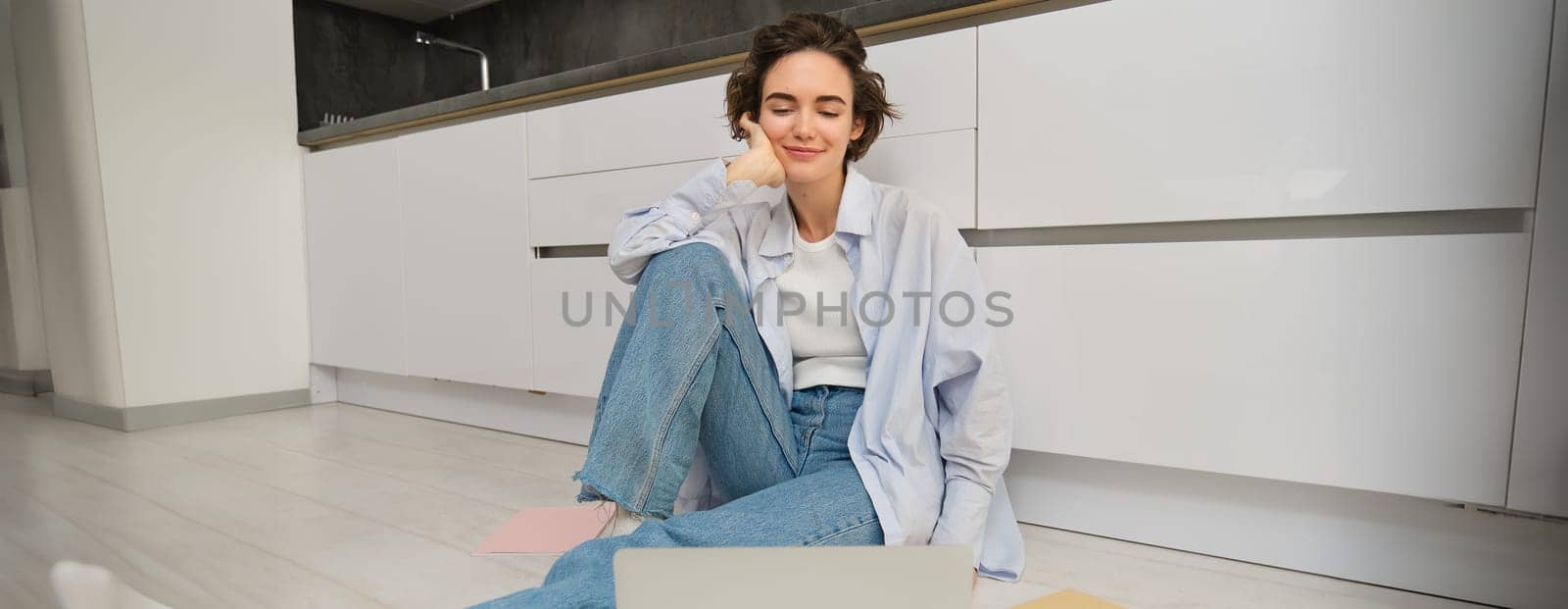 Portrait of smiling young woman watching video on laptop, sitting on kitchen floor and looking at computer screen, connects to online chat. Copy space