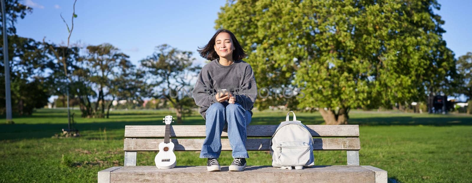Young woman with ukulele, sitting on bench in park, using mobile phone app, holding smartphone and smiling by Benzoix