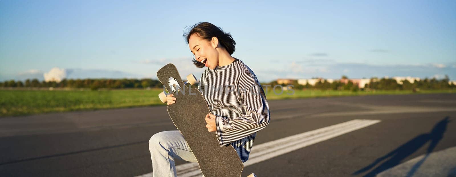 Beautiful asian teen girl playing with her longboard, holding skateboard as if playing guitar, standing on road on sunny day.