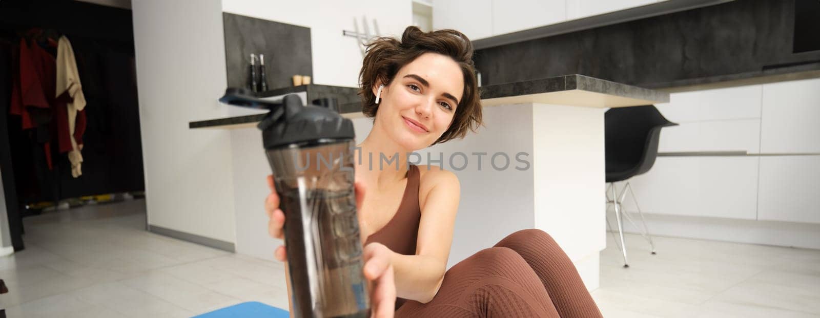 Smiling brunette woman sits on yoga fitness mat, gives you water bottle to drink after workout, does pilates at home in kitchen, looks happy at camera.