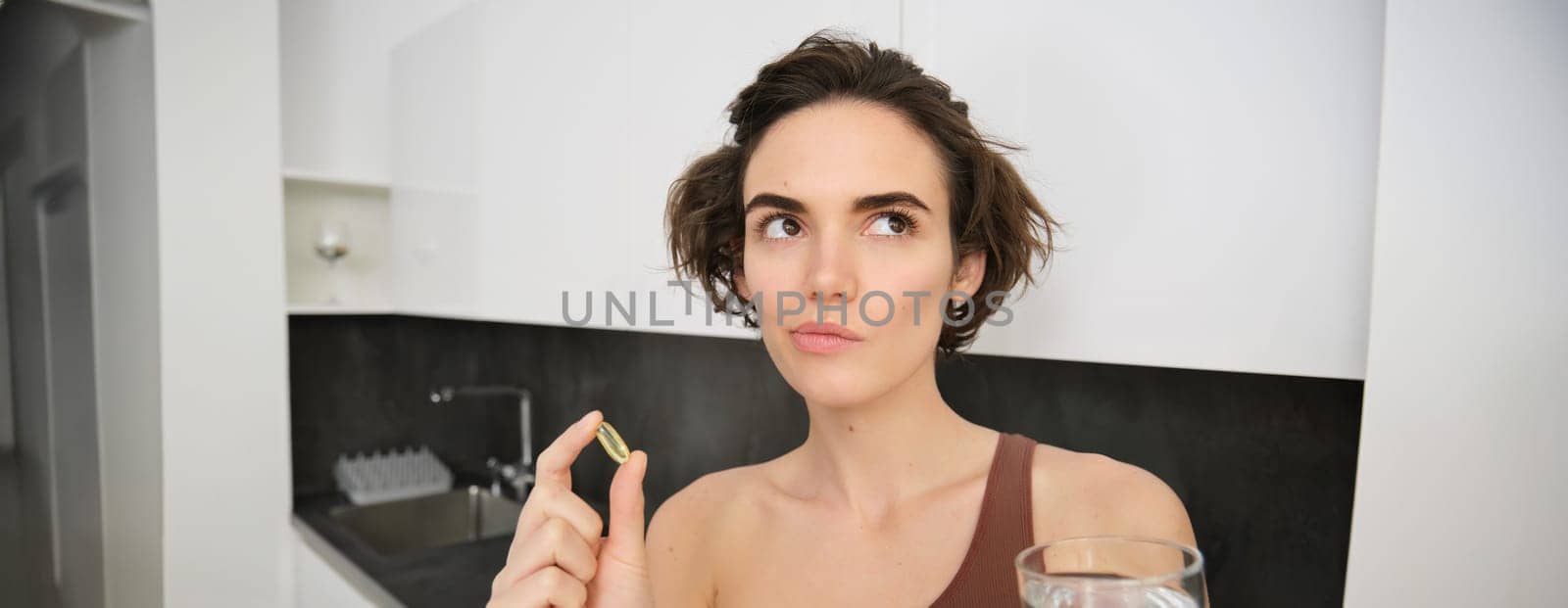 Close up portrait of sportswoman in activewear, holding omega-3, vitamin c, d and b in her hand, taking dietary supplement with drinking glass of water. Healthy lifestyle and sport concept.