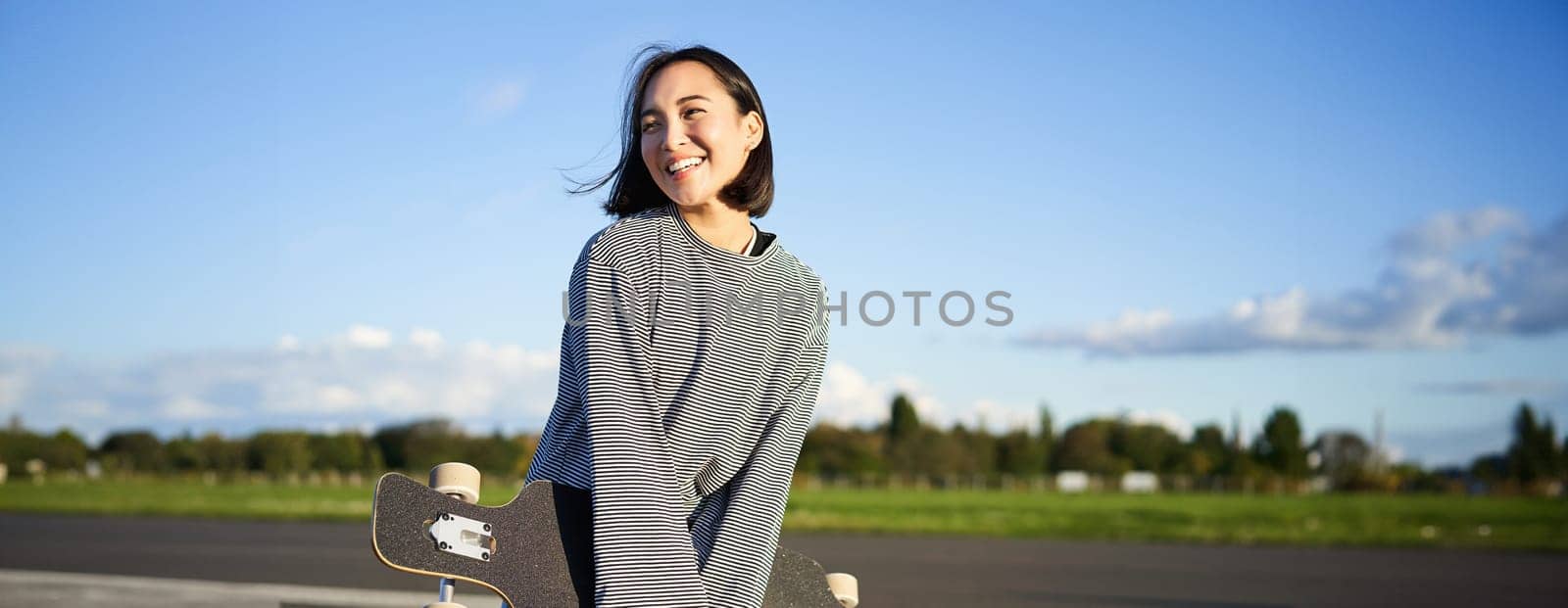 Portrait of asian woman with longboard. Korean girl skating, holding skateboard in hands, posing on road, smiling and looking aside.
