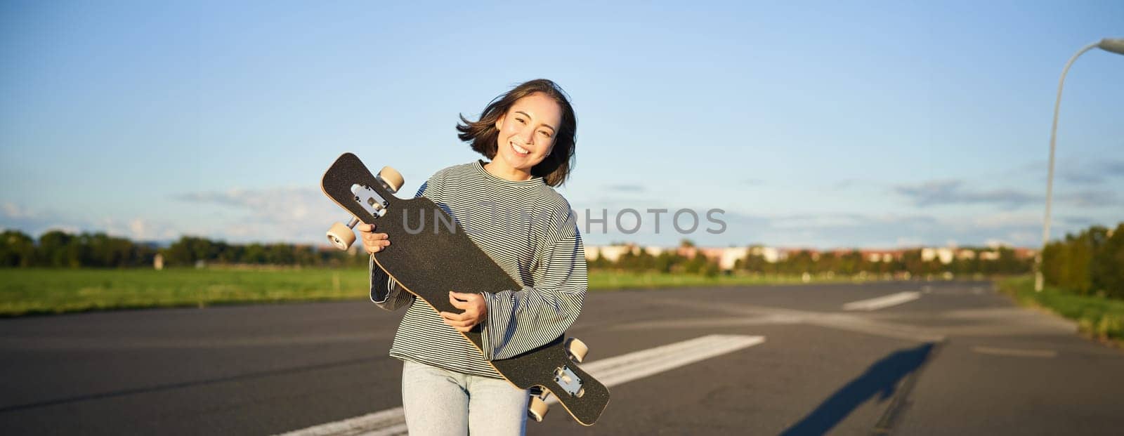 Lifestyle and hobbies. Smiling cute asian girl holding skateboard and walking towards sun on an empty road by Benzoix