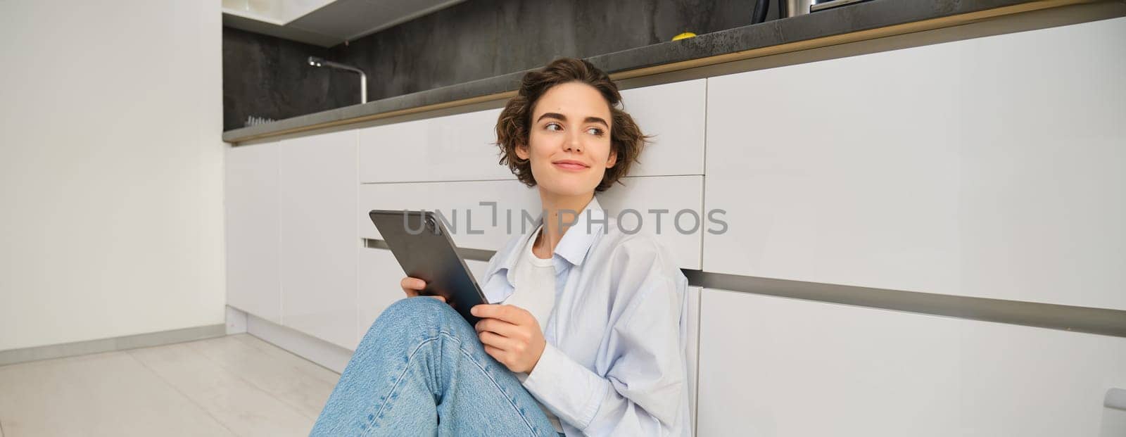 Image of happy young woman sits at home on floor, holds digital tablet, does online shopping, reading e-book. Copy space