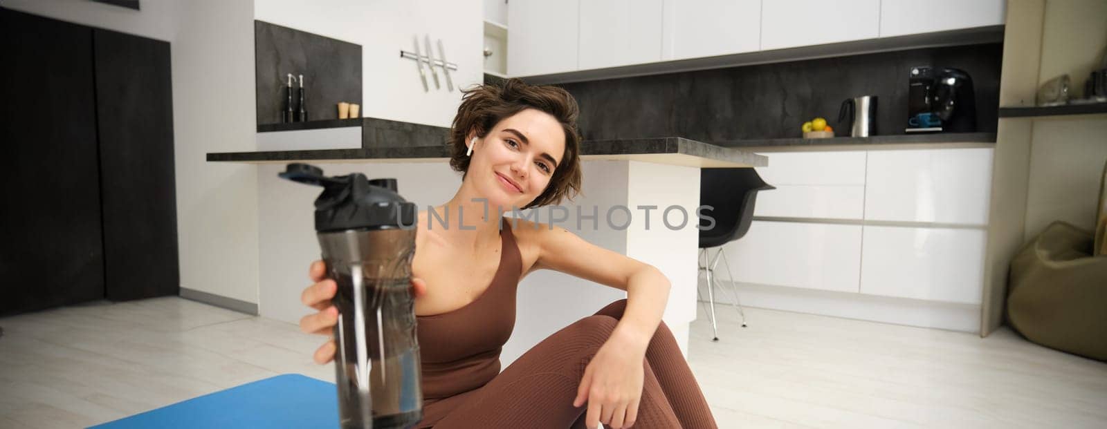 Smiling sportsgirl, woman after fitness training, gives you water bottle, stays hydrated after workout, sits on fitness yoga mat at home on floor by Benzoix