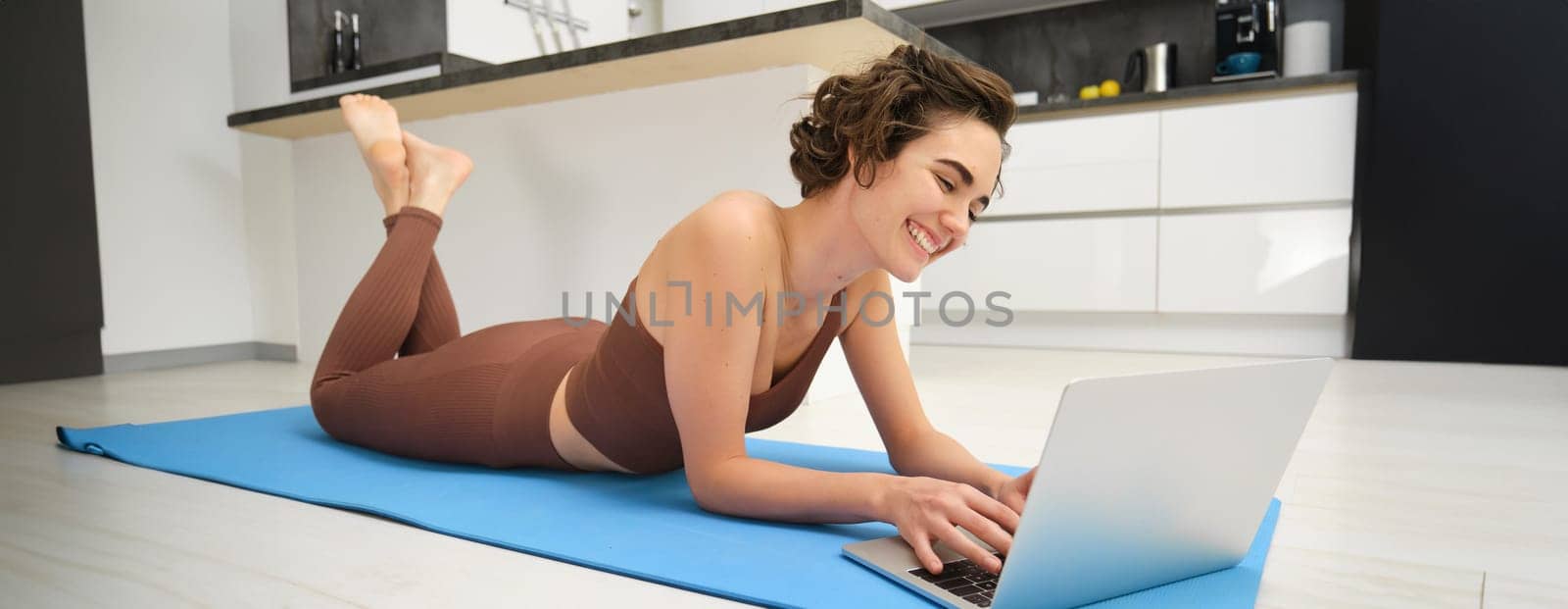 Portrait of beautiful sportswoman, fitness girl watching videos on laptop during workout, follow online pilates videos while exercise at home, wearing activewear in kitchen by Benzoix