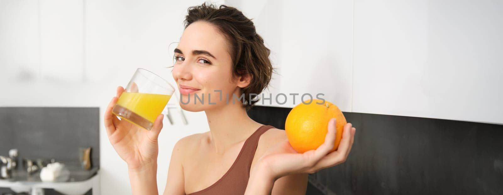 Image of sportswoman, fitness girl holding glass of juice and an orange, smiling, drinking vitamin beverage after workout, standing in her kitchen at home. Healthy lifestyle and sport concept.