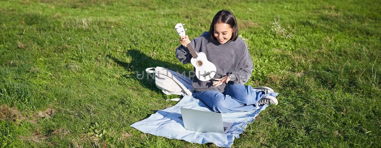 Cheerful asian girl, musician waves at her laptop, say hi to video chat, plays ukulele and records it on laptop, sits on grass in park.