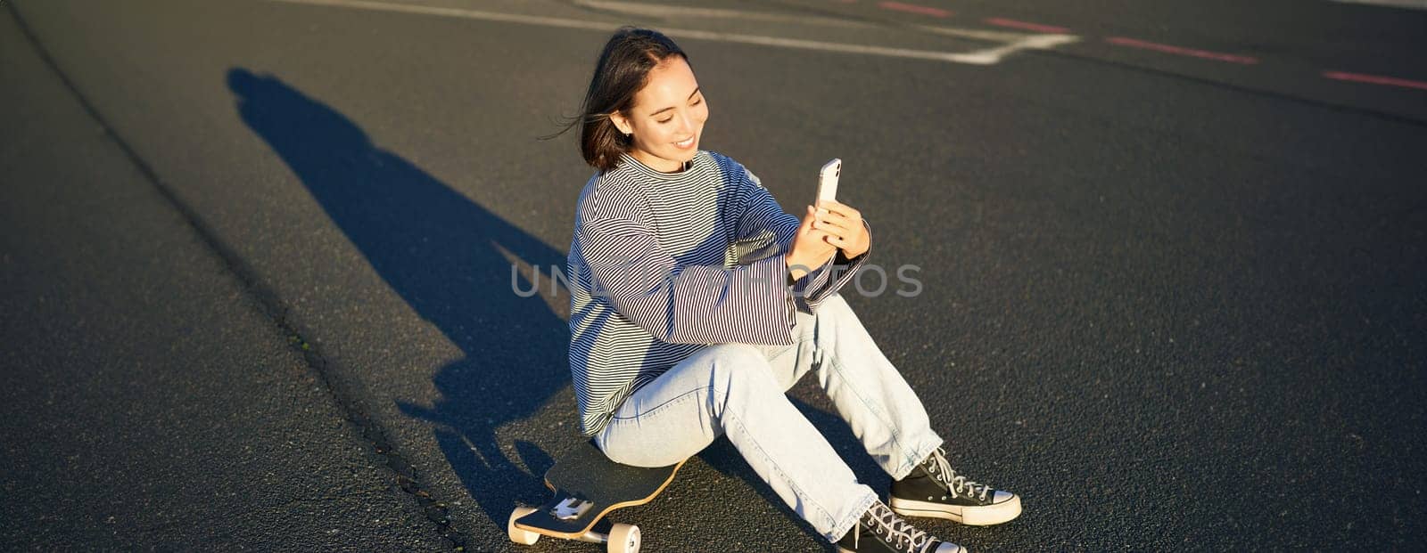 Happy asian girl sits on skateboard, takes selfie with longboard, makes cute faces, sunny day outdoors.