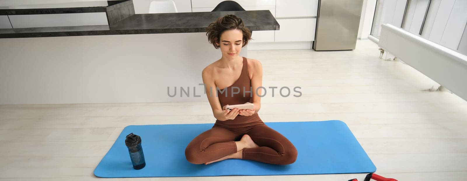 Home fitness and lifestyle. Young woman in fitness clothes, sits on sports mat at home, watching yoga tutorial on smartphone app, workout indoors.
