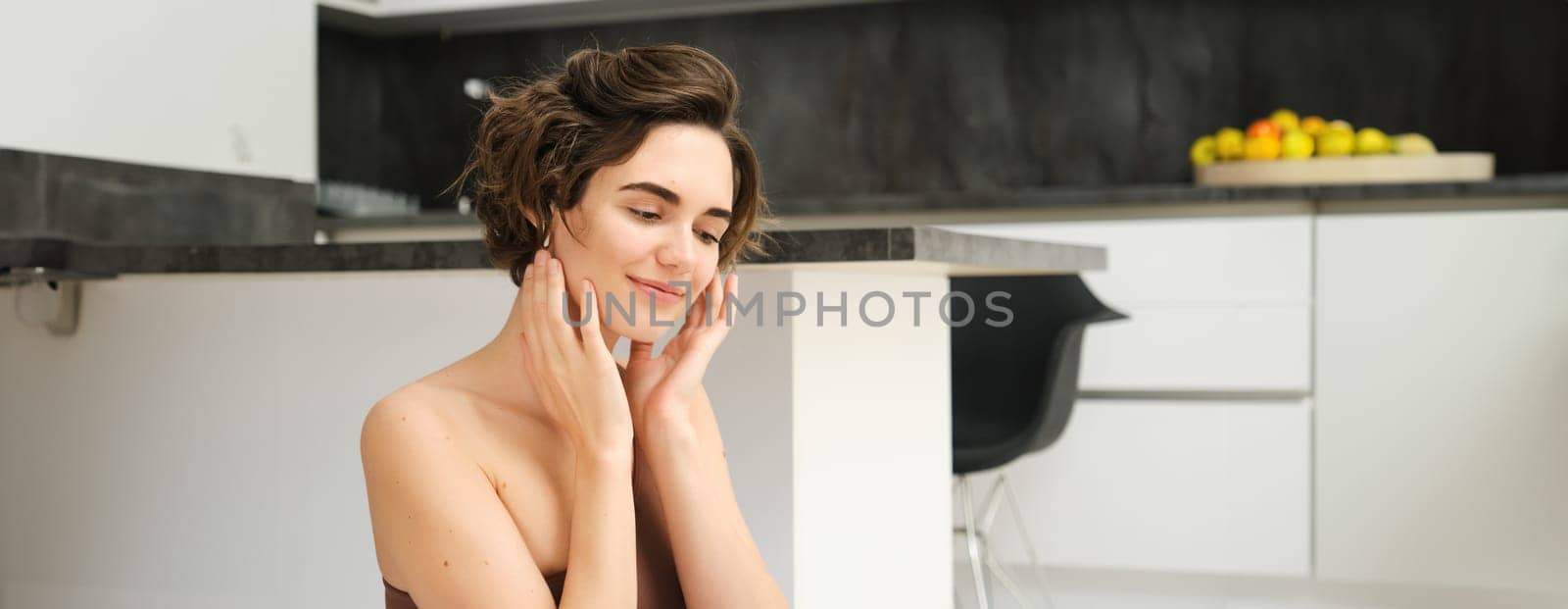 Close up of young smiling brunette woman, fitness girl in kitchen, looks aside, touches her face, listens to mindful app training session in wireless earphones, workout at home by Benzoix