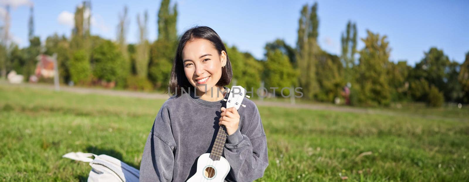 Smiling asian girl learns how to play ukulele on laptop, video chat with music teacher, sitting with instrument in park on grass by Benzoix