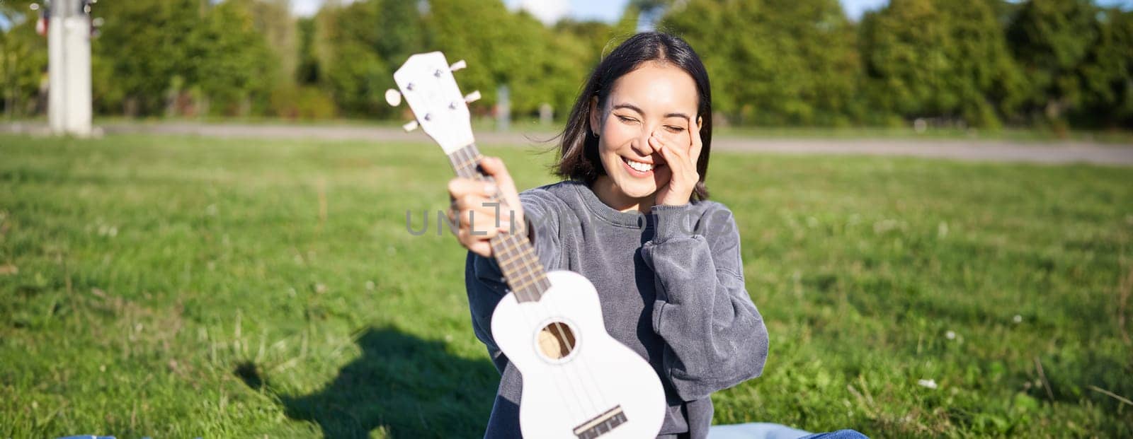 Beautiful asian girl with happy smile, shows her ukulele, sits outside in park on grass, relaxes with music.
