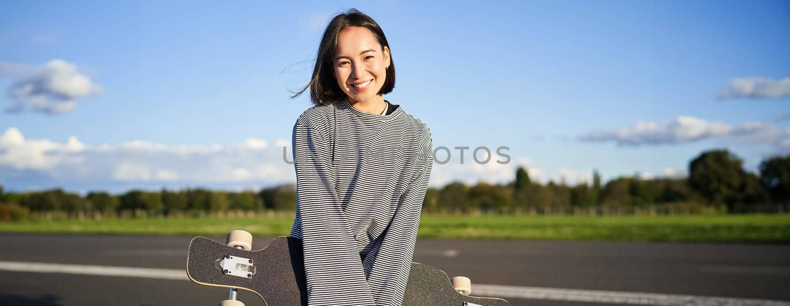 Portrait of beautiful asian girl skating on longboard, crusing with skateboard on empty road, enjoying freetime on fresh air.