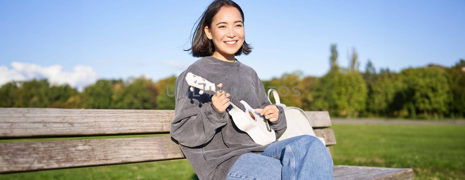 Happy cute girl sits alone on bench in park, plays ukulele guitar and enjoys sunny day outdoors by Benzoix