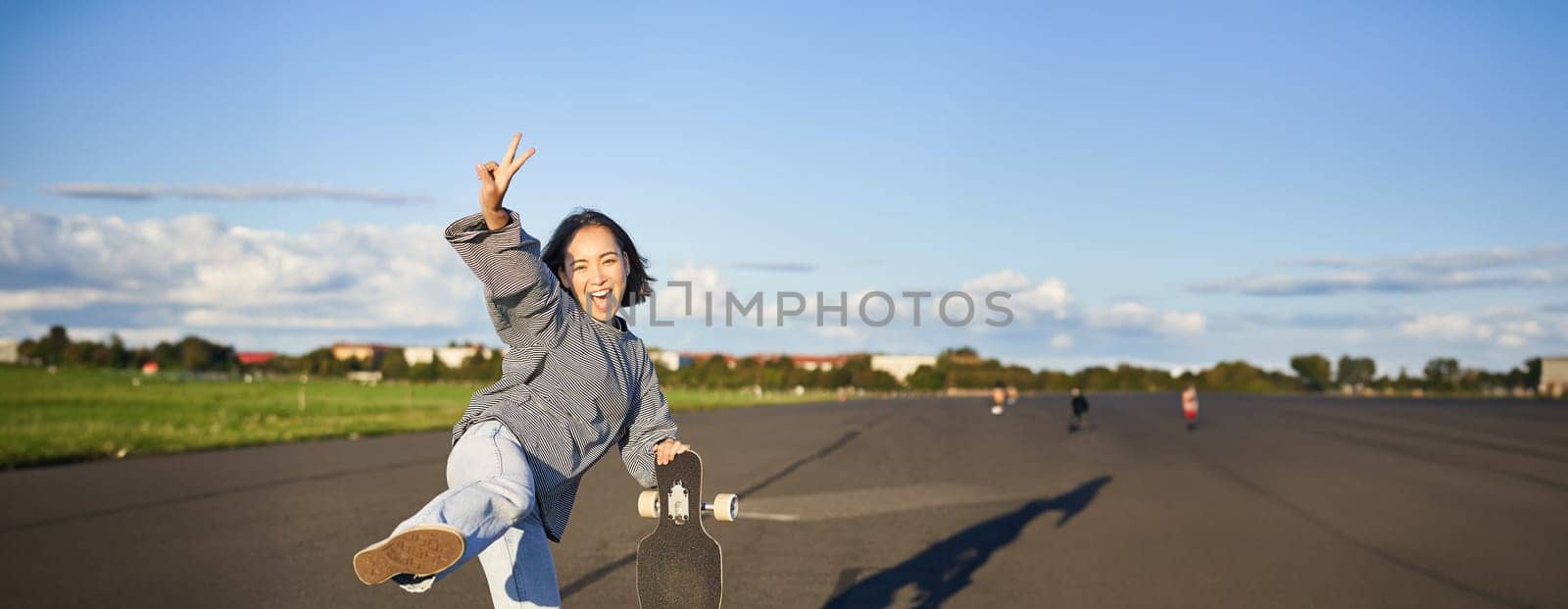 Vertical shot of happy asian skater girl, jumping, standing with skateboard and smiling. Woman skating on longboard and having fun.