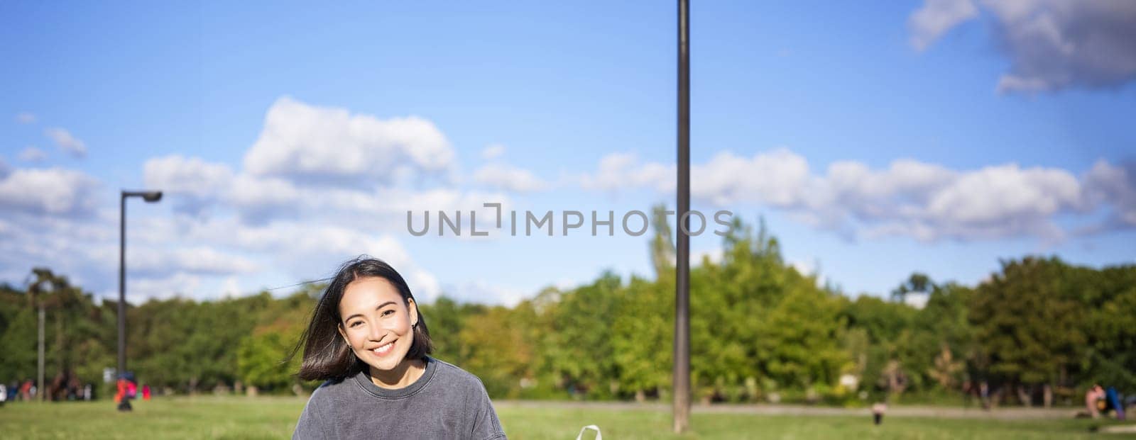 Vertical shot of cute teen girl sits in park on grass with backpack and her book, reading alone outdoors.