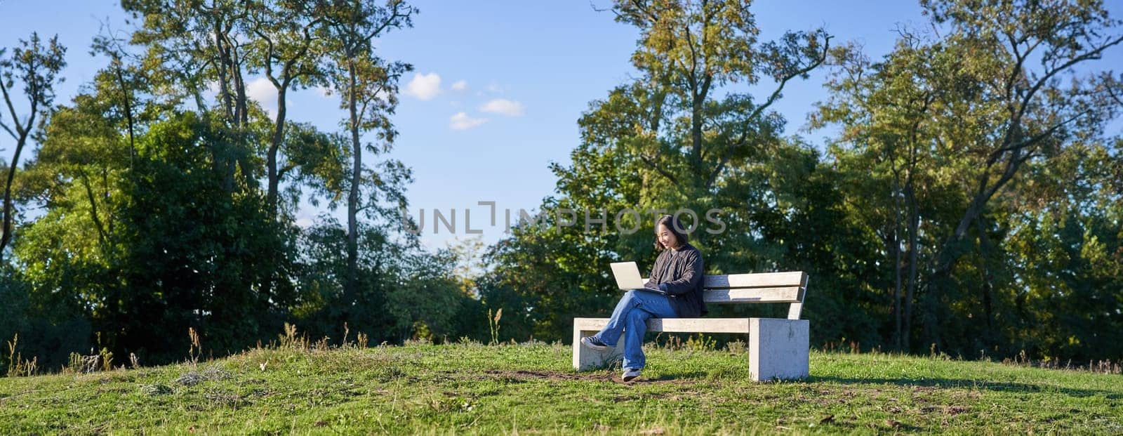 Portrait of young student, girl using laptop, sitting in park on bench, typing on computer.