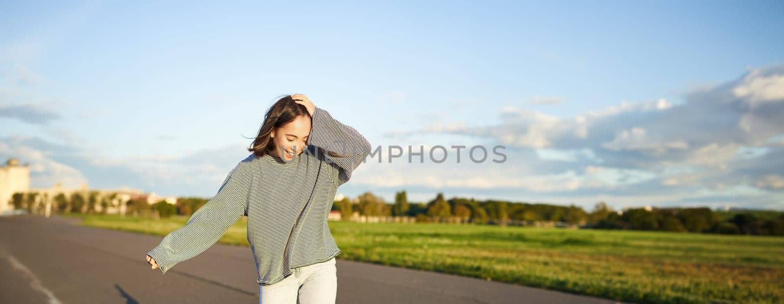 Vertical portrait of happy asian girl enjoying skateboard fun day out. Smiling korean skater on longboard, riding along empty street on sunny day.