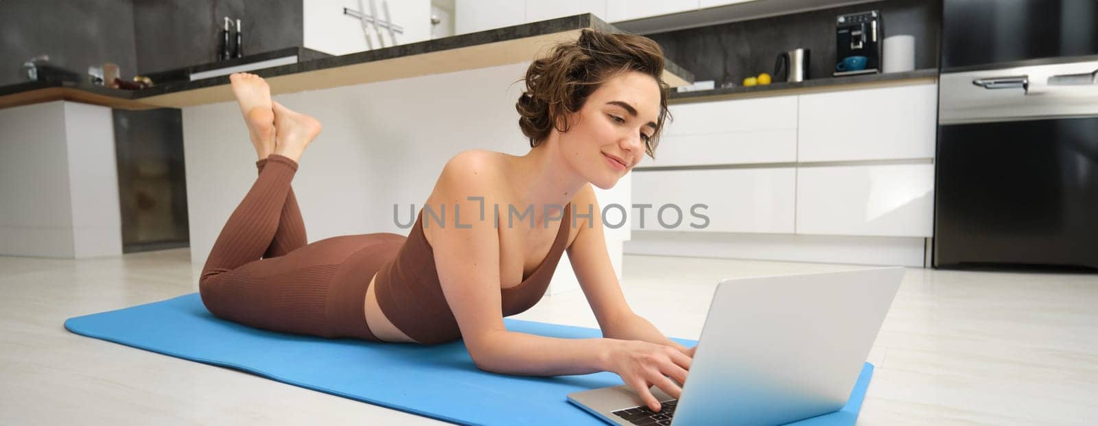 Portrait of woman athlete, girl workout at home, watches fitness videos on laptop, lying on rubber mat and doing indoor yoga session by Benzoix