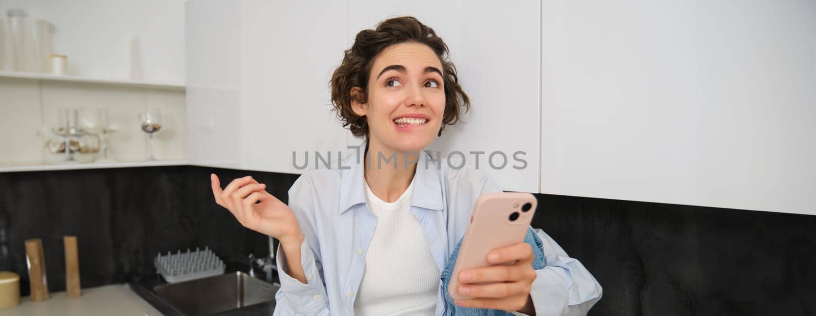 Enthusiastic girl in kitchen, holds smartphone looks excited, smiles and bites lip, tempted to try smth, talks on mobile phone.