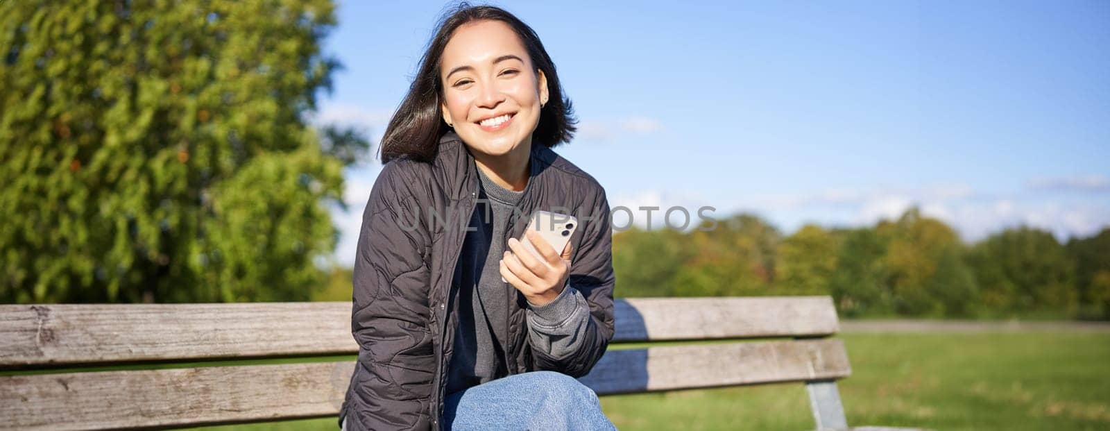 Cute young woman with smartphone in hands, sitting on bench and smiling, using mobile phone, waiting for someone in park.