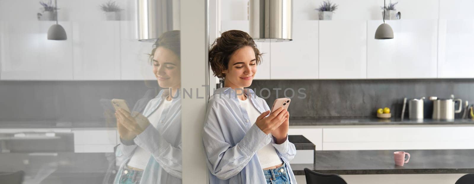 Image of young smiling woman at home, looks at her smartphone with happy face, reads message on telephone, chats on mobile phone app. Technology and communication concept