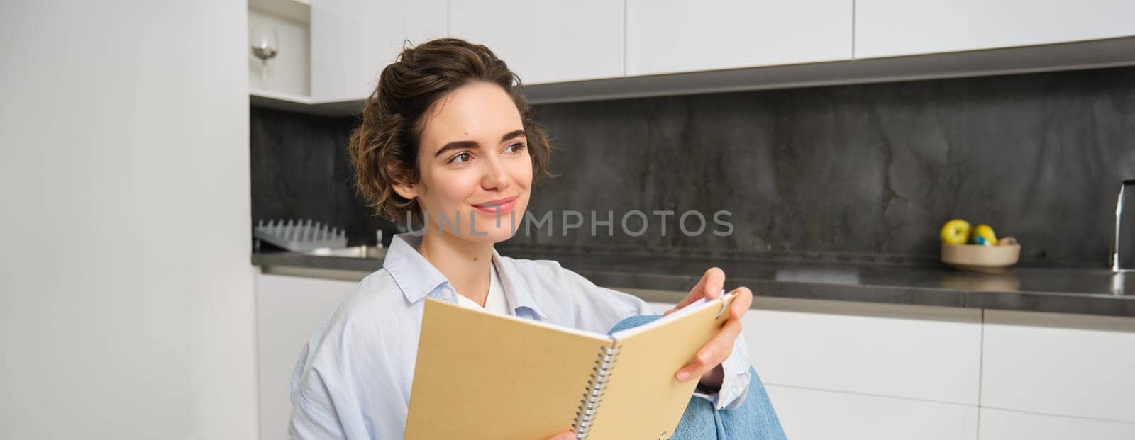 Portrait of happy smiling woman, holding notebook, studying in her kitchen, reading journal. Lifestyle and people concept