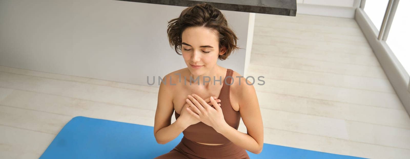 Portrait of young mindful woman, opens her chakra, doing yoga on rubber mat at home, closes eyes and smiles, feels peace inside her chest. Sport and lifestyle concept