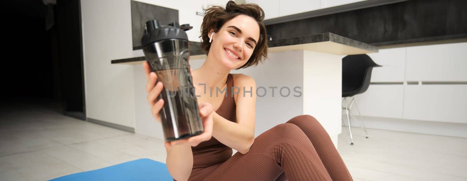 Smiling sportsgirl, woman after fitness training, gives you water bottle, stays hydrated after workout, sits on fitness yoga mat at home on floor.