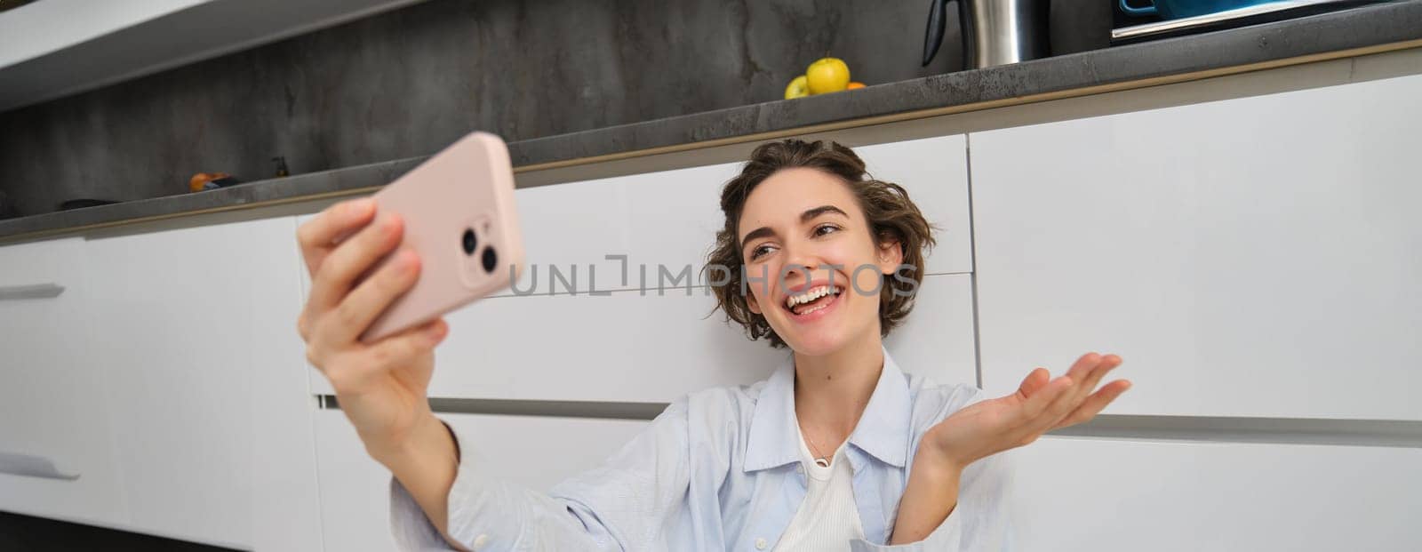 Portrait of young woman video chats, records vlog while sits on kitchen floor. Cute girl takes selfie on smartphone.