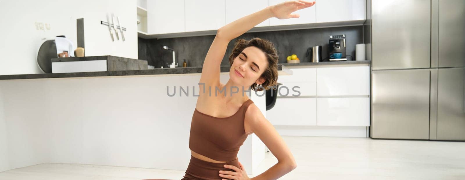 Portrait of fitness woman doing exercises in bright room at home, stretching in lotus pose, sitting on rubber yoga mat and doing workout training, drinking water from bottle.