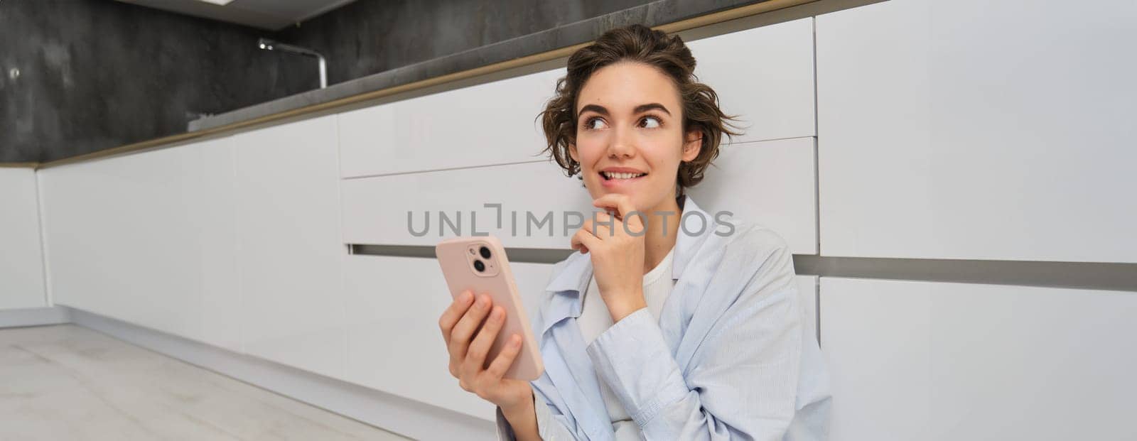 Image of smiling woman thinking, using smartphone and pondering, making decision, online shopping on mobile phone.
