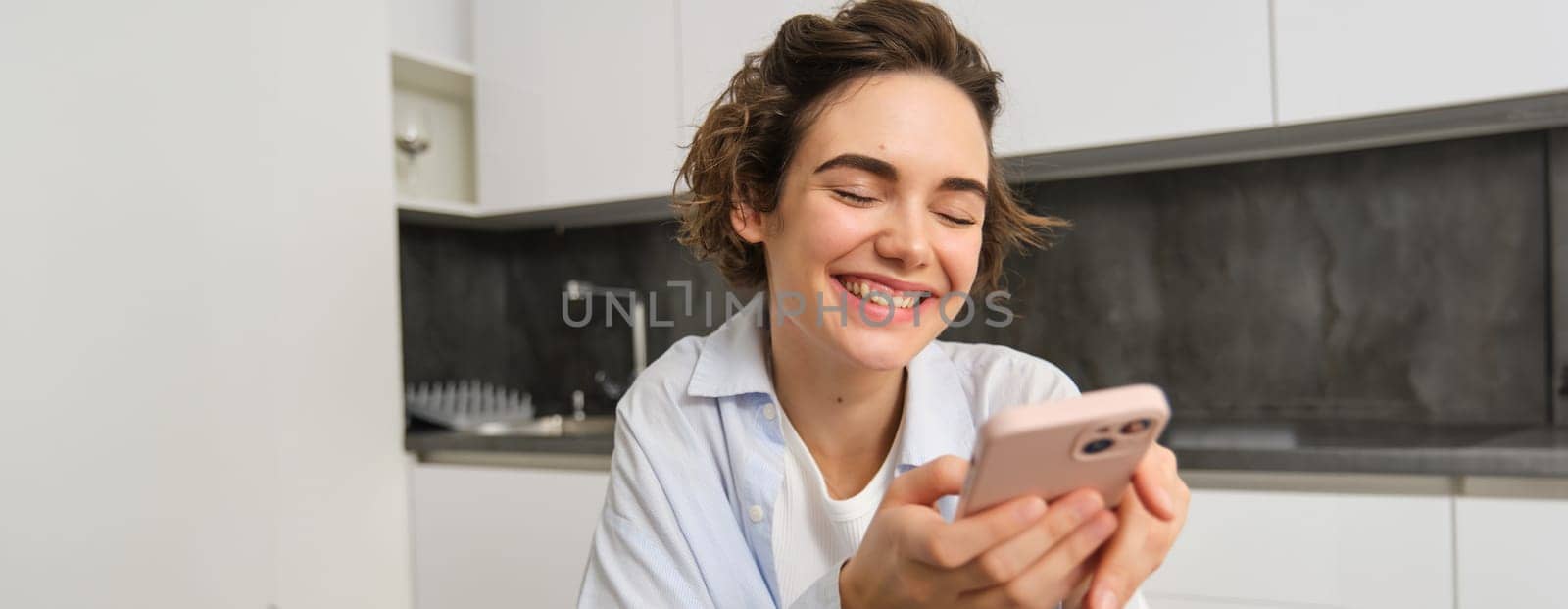 Portrait of modern brunette woman holding mobile phone and smiling. Girl sits in kitchen at home with smartphone, using app to order delivery, does online shopping.