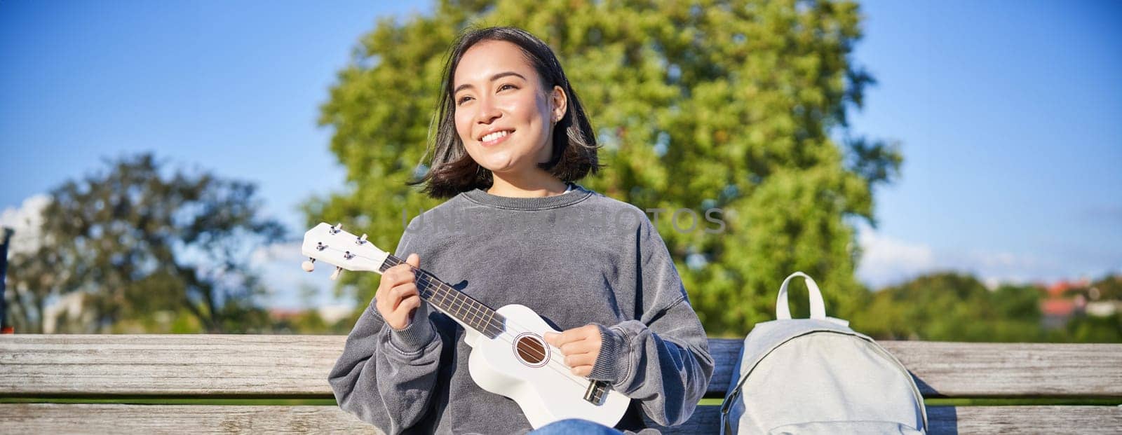 Lovely young woman sitting with backpack on bench in sunny park, plays ukulele guitar and sings along by Benzoix