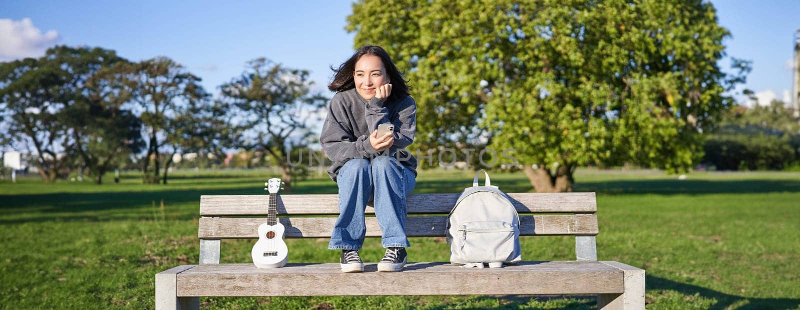 Young woman with ukulele, sitting on bench in park, using mobile phone app, holding smartphone and smiling.