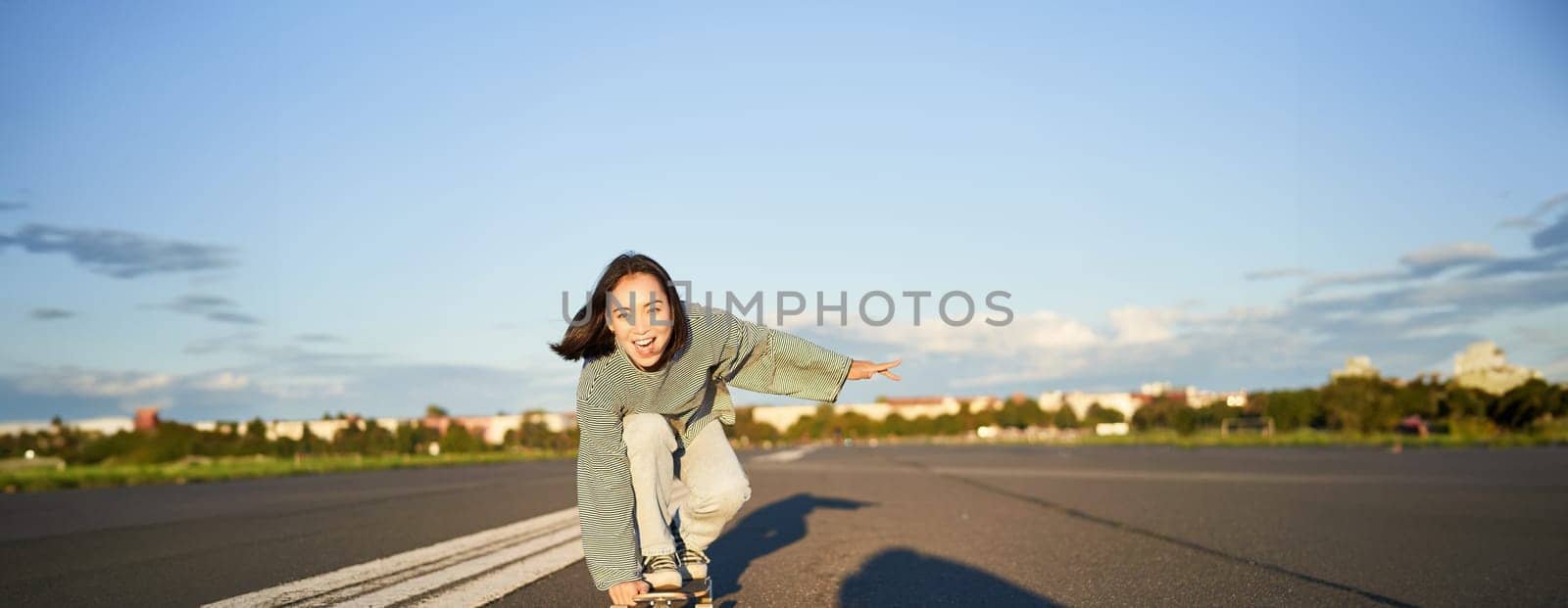 Skater girl riding on skateboard, standing on her longboard and laughing, riding cruiser on an empty street towards the sun.