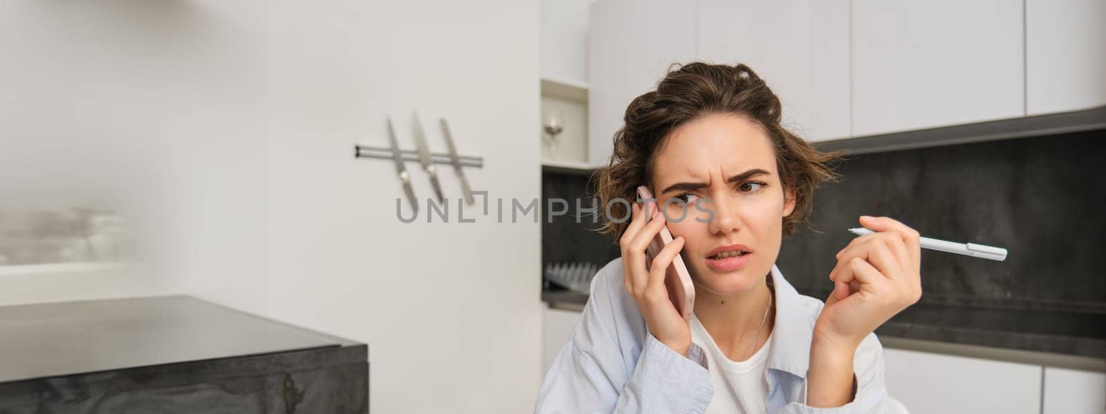Portrait of woman having complicated conversation, holding smartphone, frowning with frustrated, confused face, holding pen.