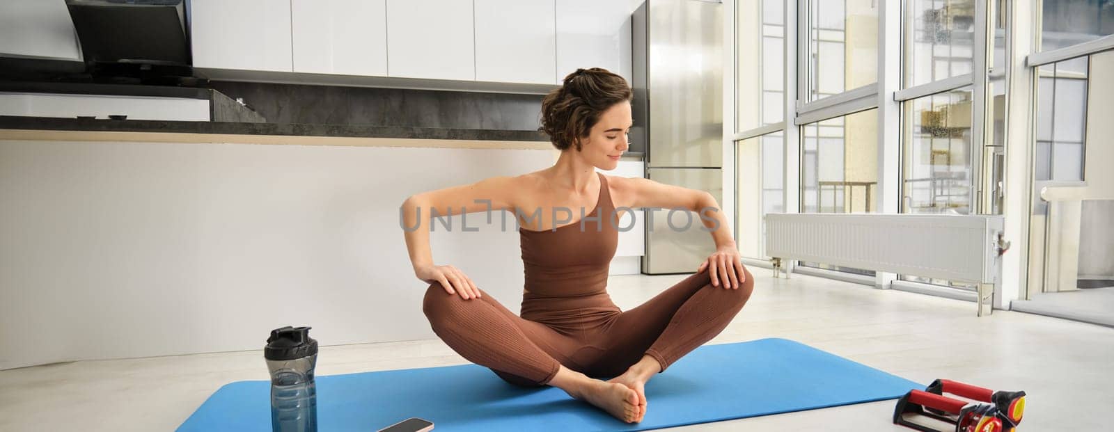 Sport and wellbeing. Young brunette fitness girl, wearing activewear, sitting on yoga mat, meditating, practice relaxation and mindfulness at home, workout in kitchen. Copy space