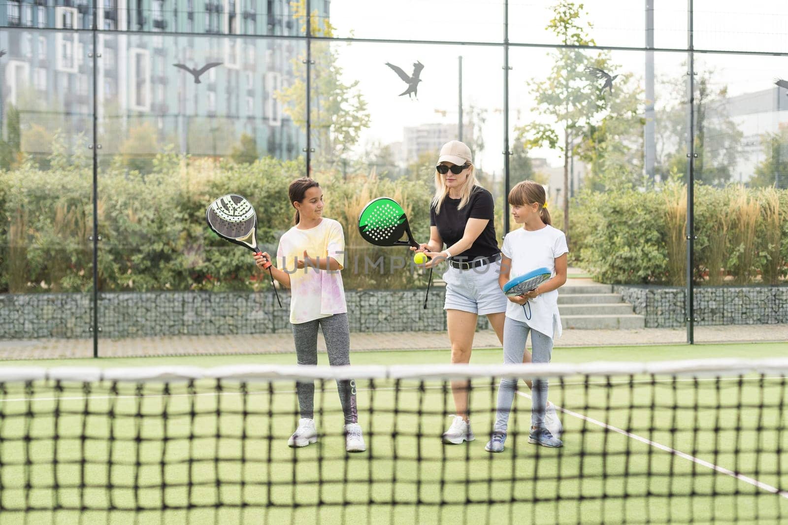 Young tennis trainer in activewear holding racket held by one of two cute girls while consulting her during individual training on stadium. High quality photo