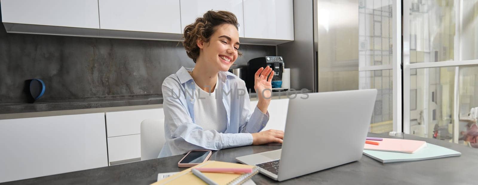 Happy young woman, tutor teacher students online. Girl connects to remote work meeting from her home laptop, says hello, waves hand at computer by Benzoix