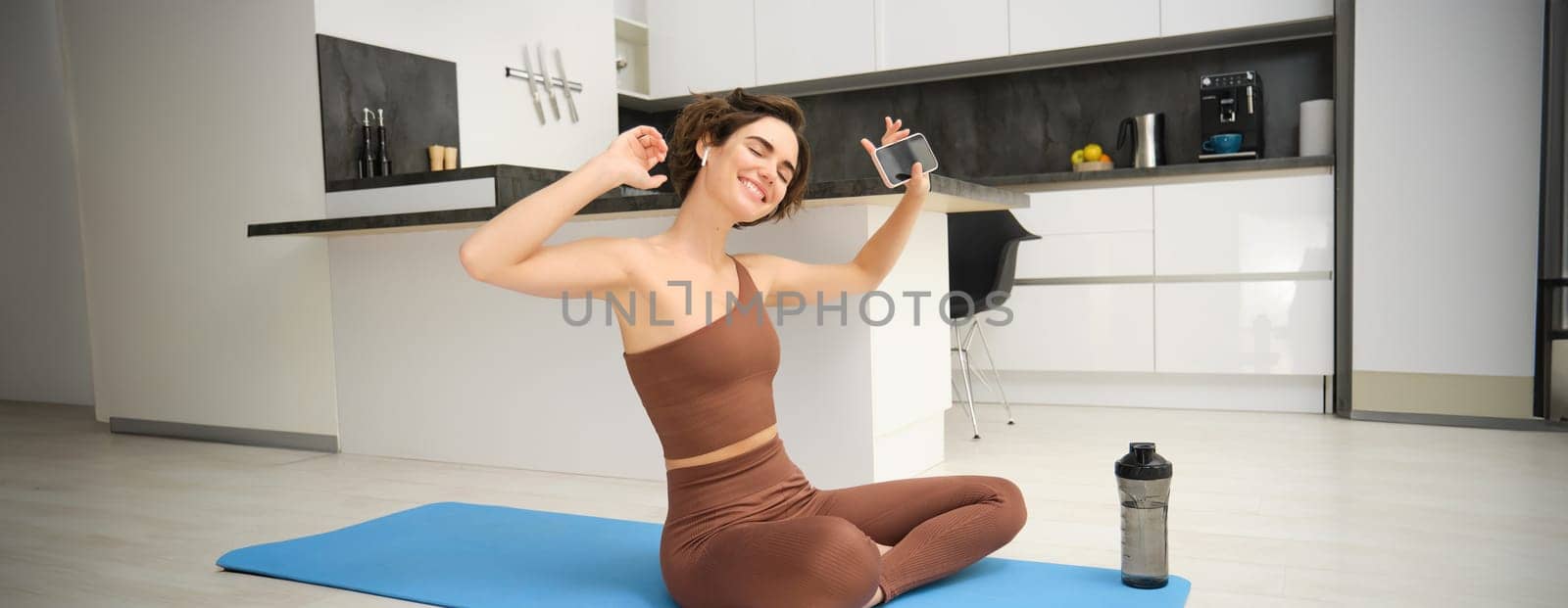 Active lifestyle and health. Happy young woman does workout, sits and dances with smartphone in hands, listens music in wireless earphones during fitness training session, yoga on floor mat.