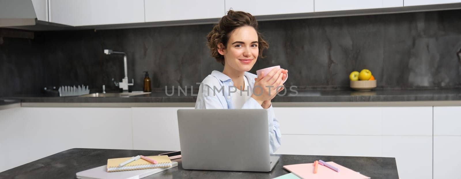 Image of young woman drinks cup of coffee and works from home. Girl browsing website, shops online on laptop and enjoys tea at home by Benzoix