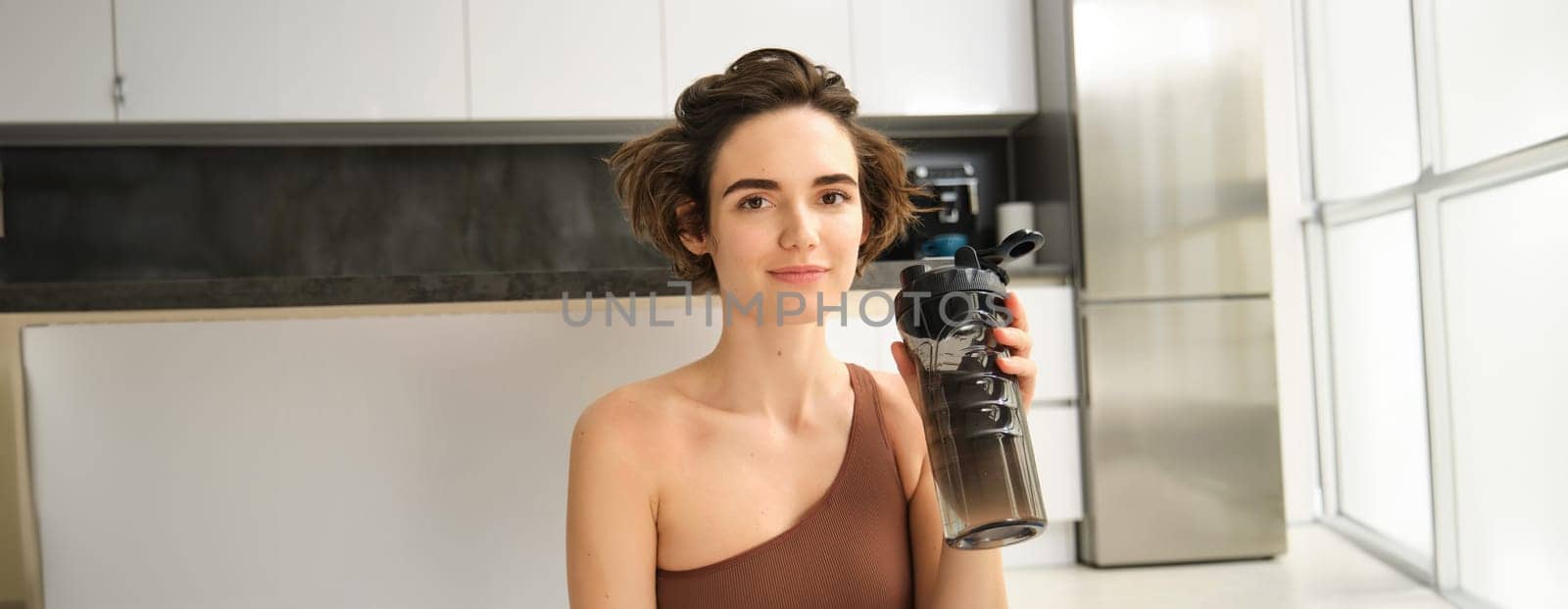 Fitness instructor, smiling young woman workout at home, sits on yoga mat and drinks water, does exercises, training session in her kitchen. Sport indoors concept