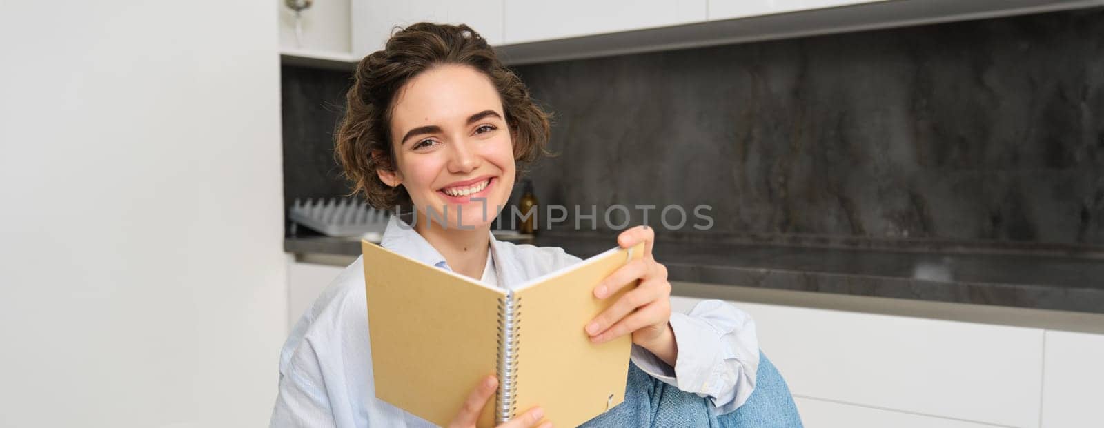 Image of beautiful girl at home, holding notebook, doing her homework, studying for exam in her kitchen and smiling by Benzoix