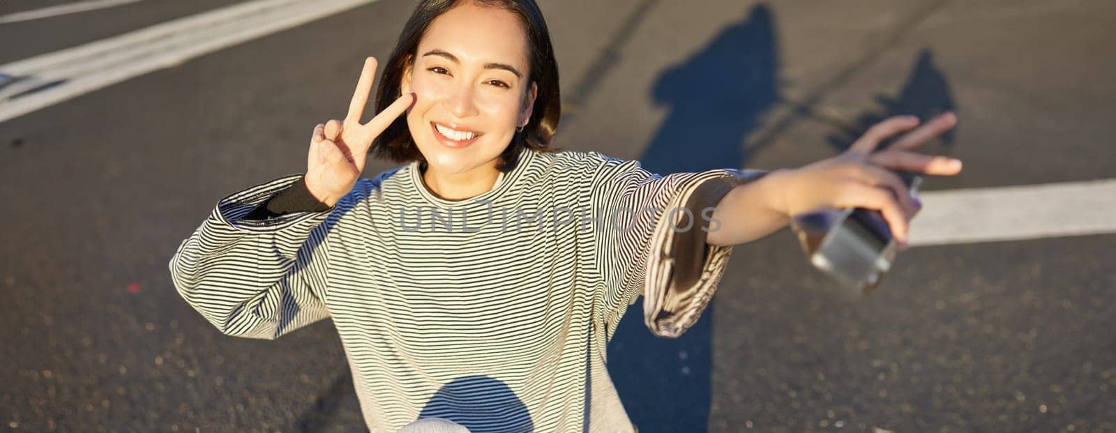 Selfie of asian girl sitting on skateboard, taking photo on smartphone, smiling and showing peace v-sign.