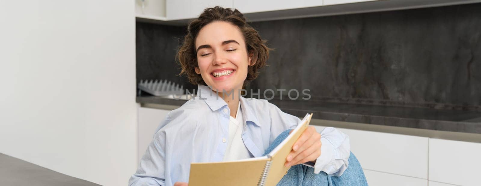 Portrait of happy smiling woman, holding notebook, studying in her kitchen, reading journal by Benzoix
