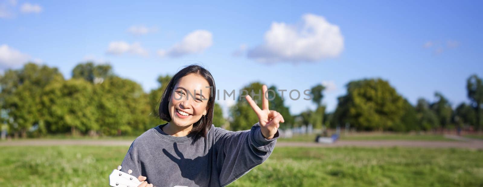 Vertical shot of positive asian girl shows peace sign, plays ukulele in park, rests and enjoys the day. Copy space.