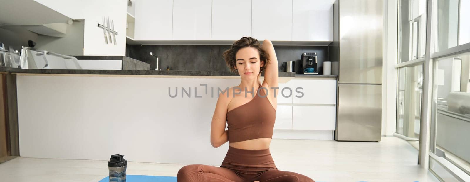 Portrait of brunette girl doing sports at home, sitting on yoga mat in bright room at home, stretching hands behind her back, workout indoors. Sport and wellbeing