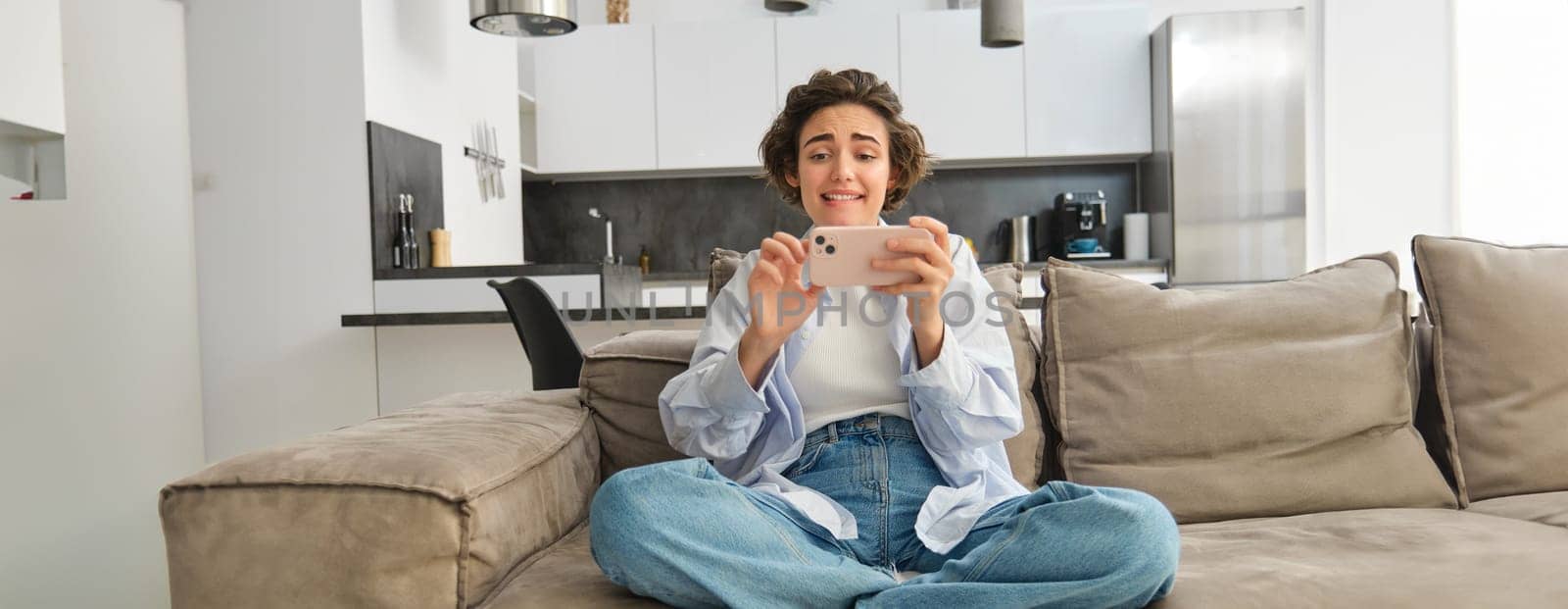 Portrait of woman playing mobile phone games, watching video with excited face, using smartphone for leisure, sits on sofa at home. Lifestyle and people concept